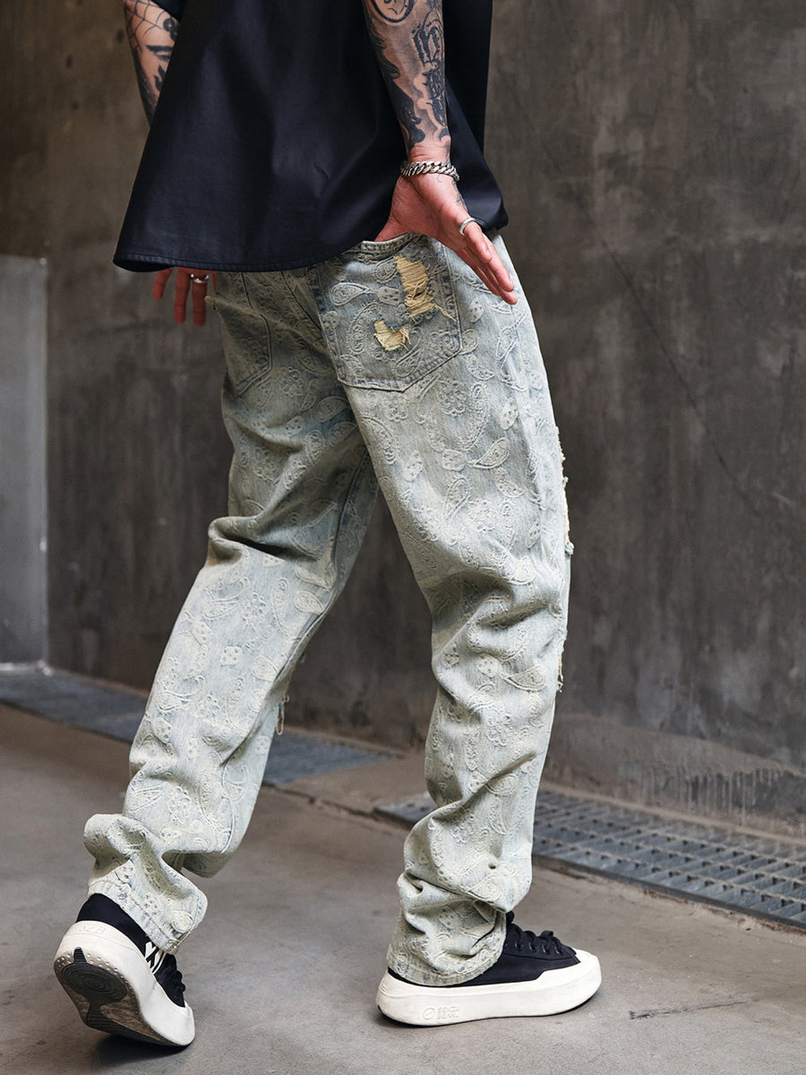 Distressed light wash men's wide-leg jeans with paisley pattern and ripped details.