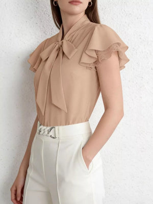 Beige tie-neck blouse with ruffle sleeves and lace trim, styled with white pants.