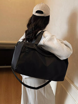 Black nylon duffel bag with adjustable strap and small logo detail, worn by a person in a grey hoodie and light pants.