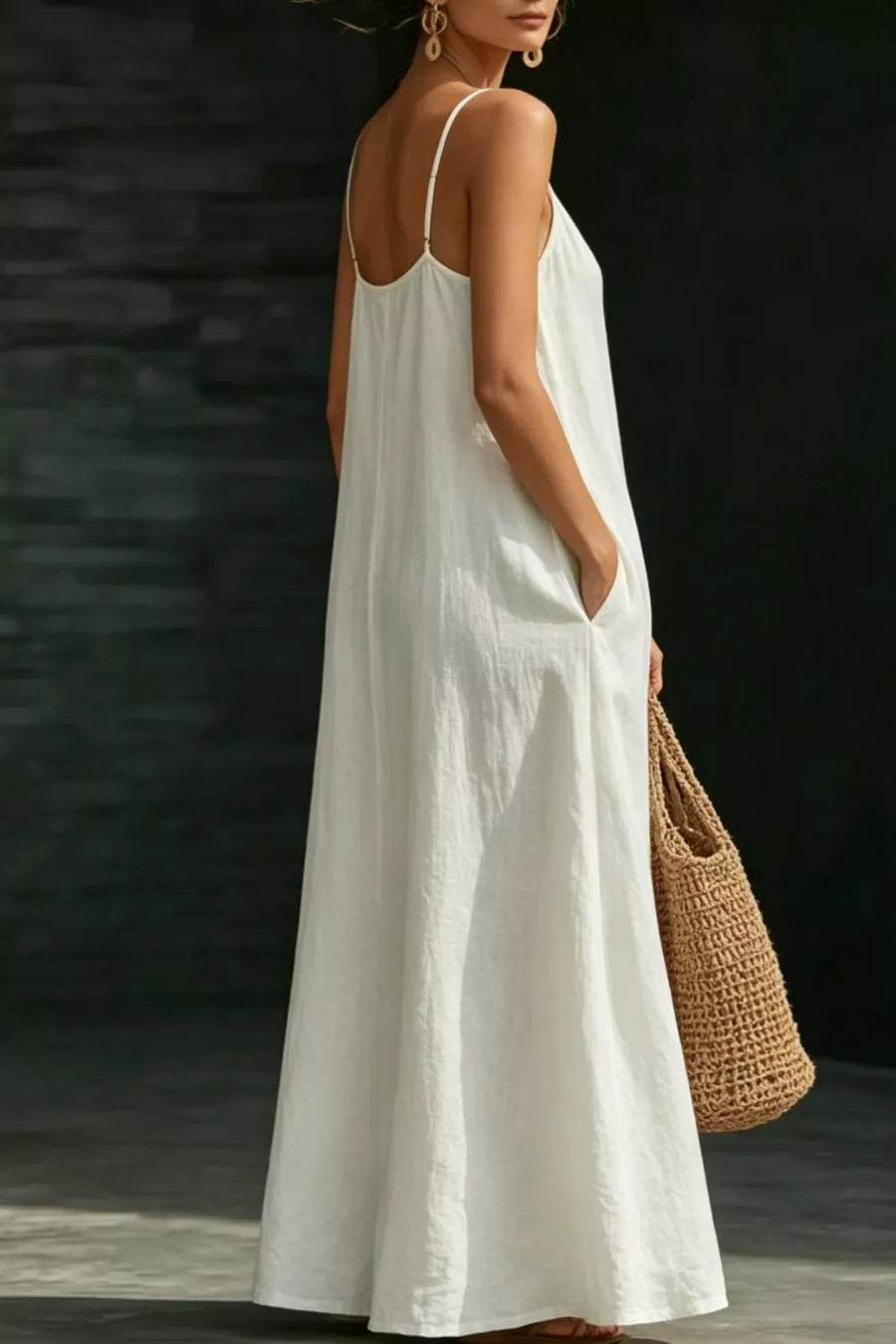 White a-line maxi dress with spaghetti straps and pockets, styled with a straw bag.
