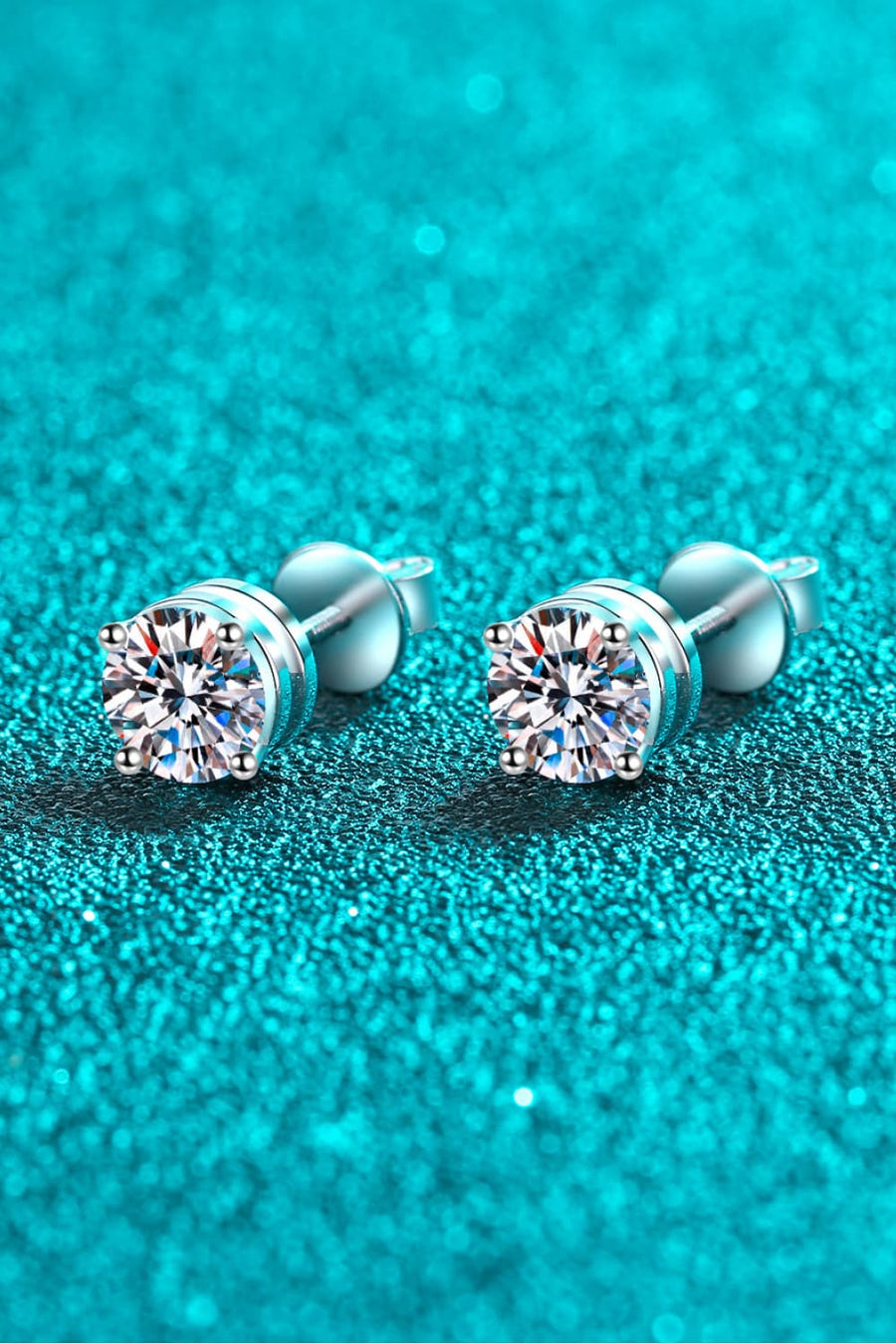 Moissanite solitaire stud earrings with rhodium finish on a teal glitter background.