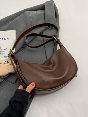 Brown crescent shoulder bag with plush charm and adjustable strap.