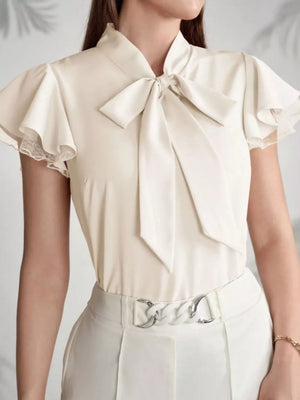 Beige tie-neck blouse with ruffle sleeves and lace trim, styled with white pants.