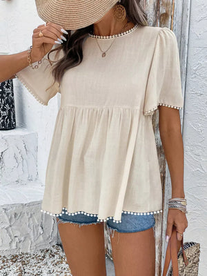 Charming babydoll blouse in cream with pom-pom trim and short sleeves, styled with denim shorts.