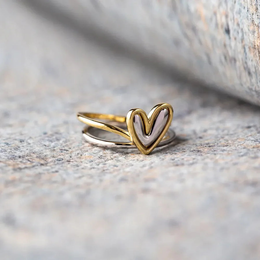 Two-tone heart outline ring in gold and silver, stacked on a stone surface.