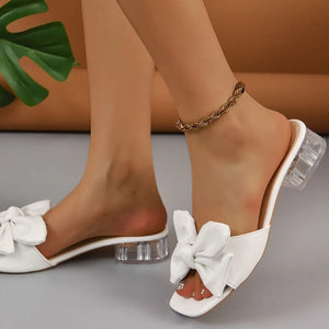 White bow slides with clear block heels and a gold chain anklet.