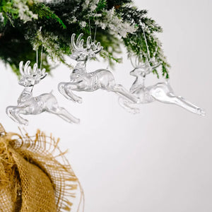 Clear acrylic reindeer Christmas ornaments hang from a snowy pine branch.