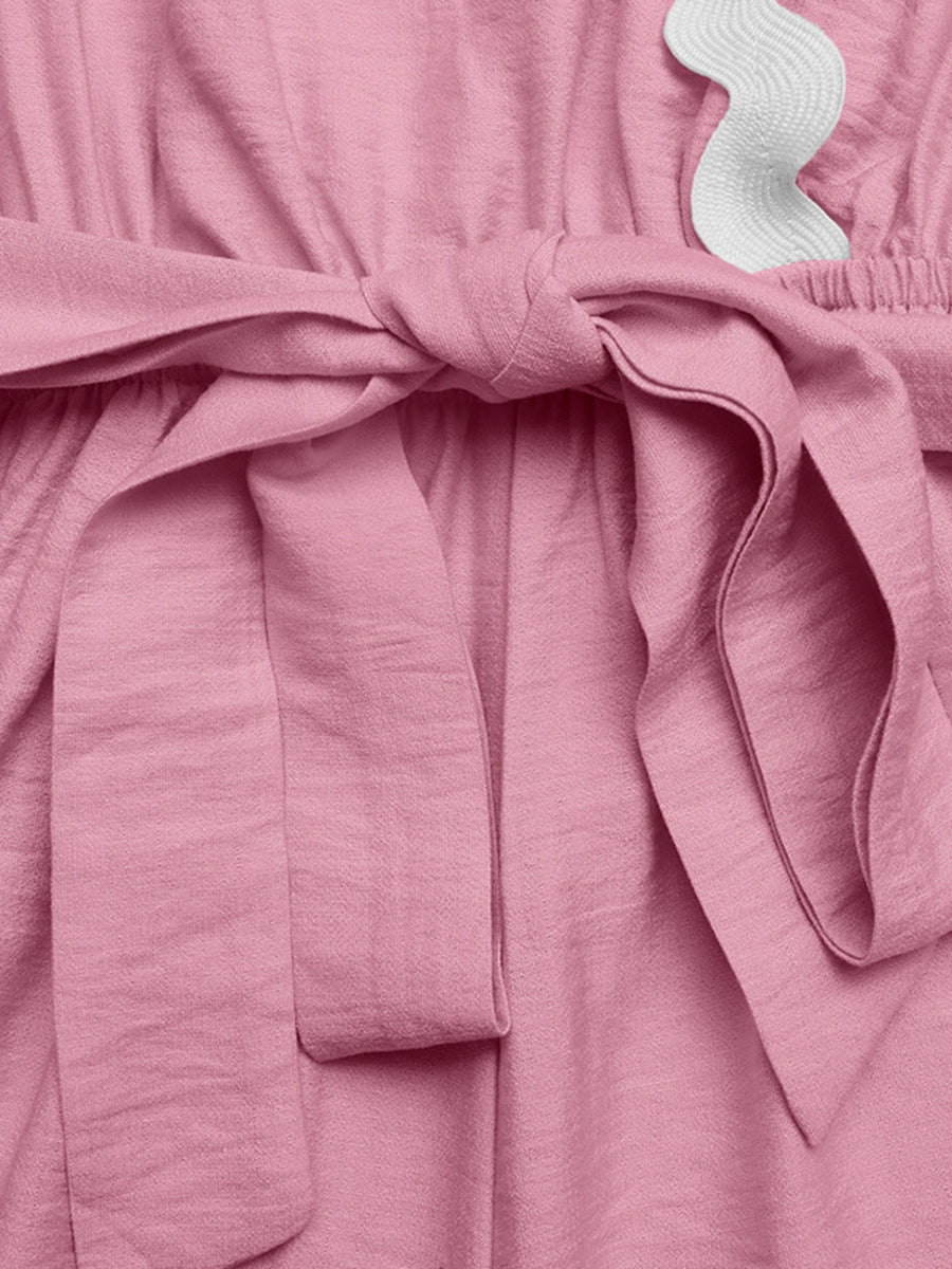 Pink maternity wrap romper with tie waist and white scalloped trim detail.