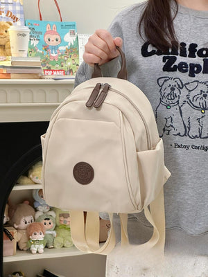 Cream nylon mini backpack with zippered pockets and a bear patch detail.