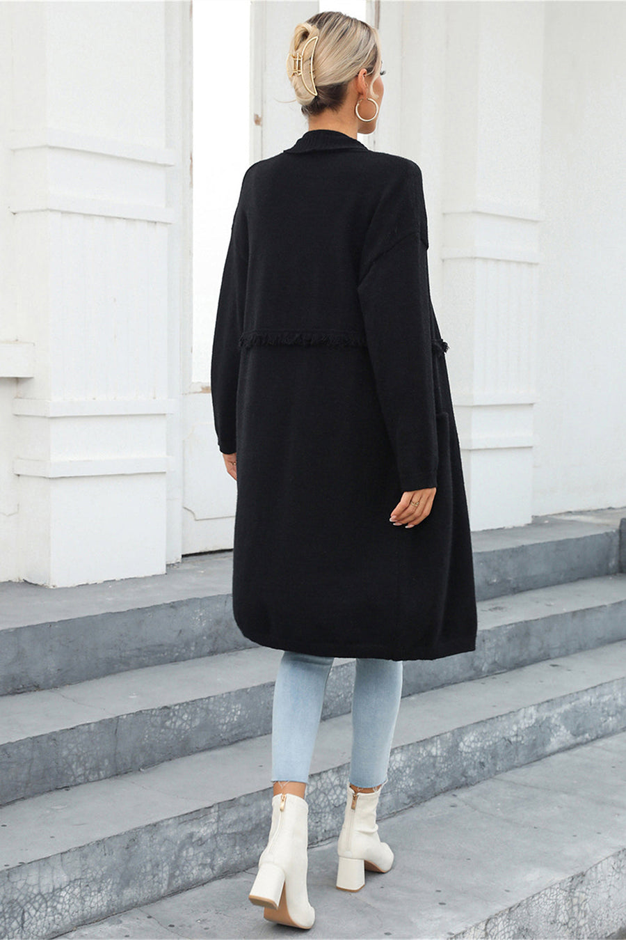 Black longline knit cardigan with fringe trim detail and pockets, worn open.