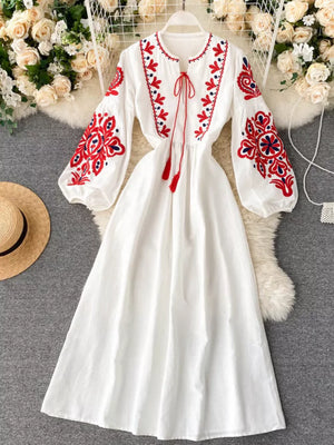 White maxi dress with red and blue embroidery, balloon sleeves, and tassel tie neckline.