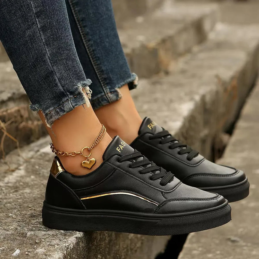 Black sneakers with gold metallic trim and heel accent, featuring a heart charm anklet.