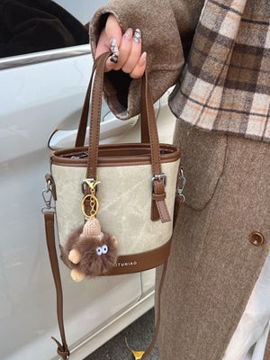 Two-tone bucket crossbody bag in beige canvas with brown trim and a fuzzy charm.