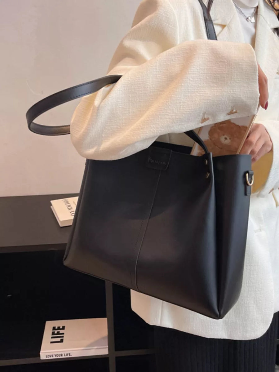 Black trapezoid PU leather handbag with wide straps and detachable strap detail.