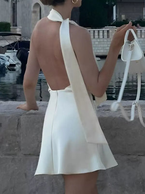 White halter neck mini dress with open back and flowing sash, styled with a white handbag.