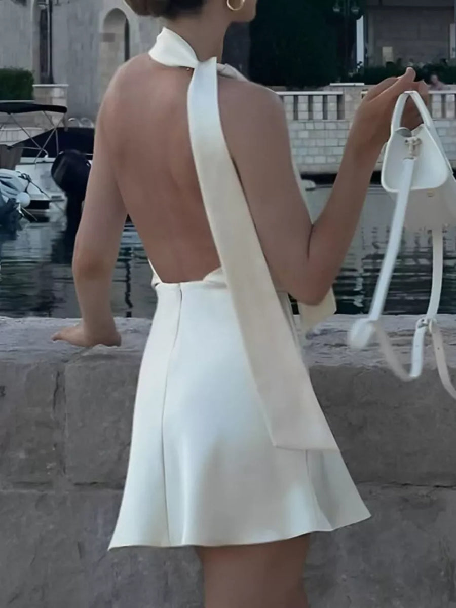 White halter neck mini dress with open back and flowing sash, styled with a white handbag.