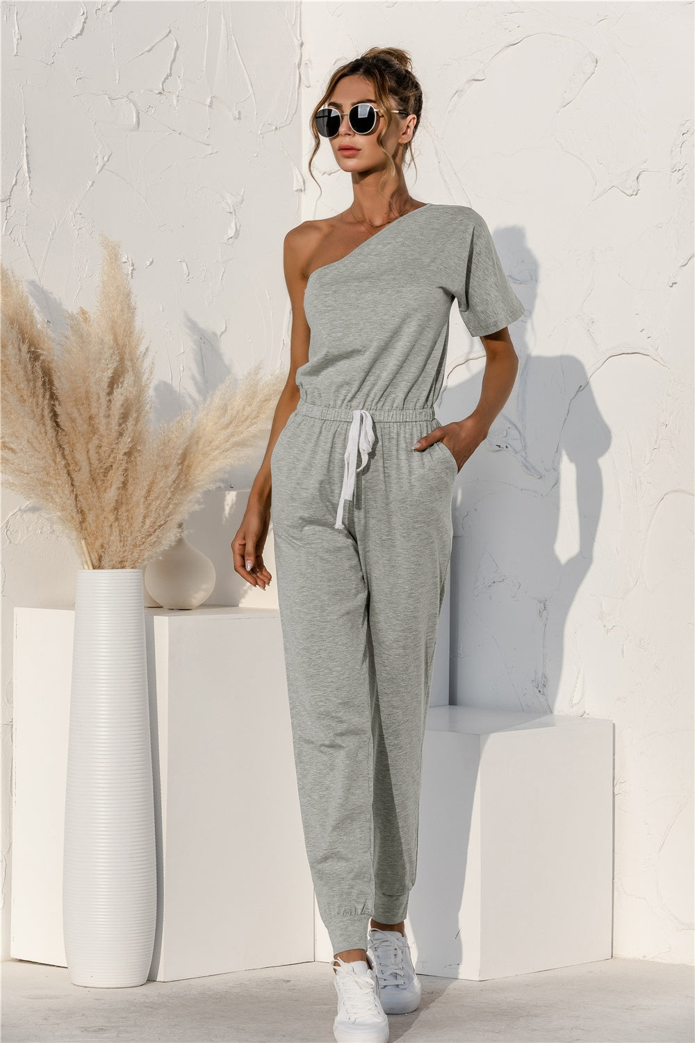 Gray asymmetric one-shoulder sweat jumpsuit with drawstring waist and cuffed jogger legs.