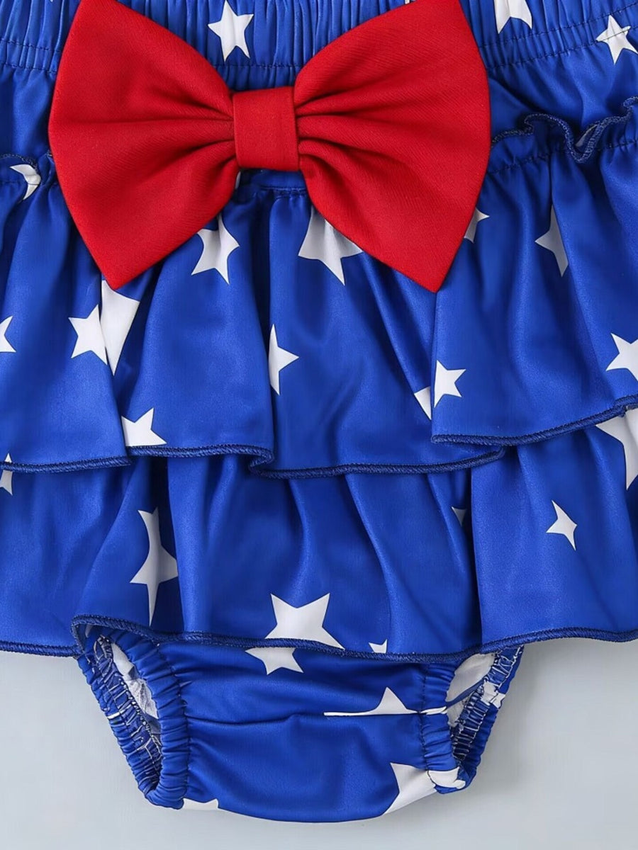 Baby romper in royal blue with white stars, tiered ruffles, and a red bow detail.