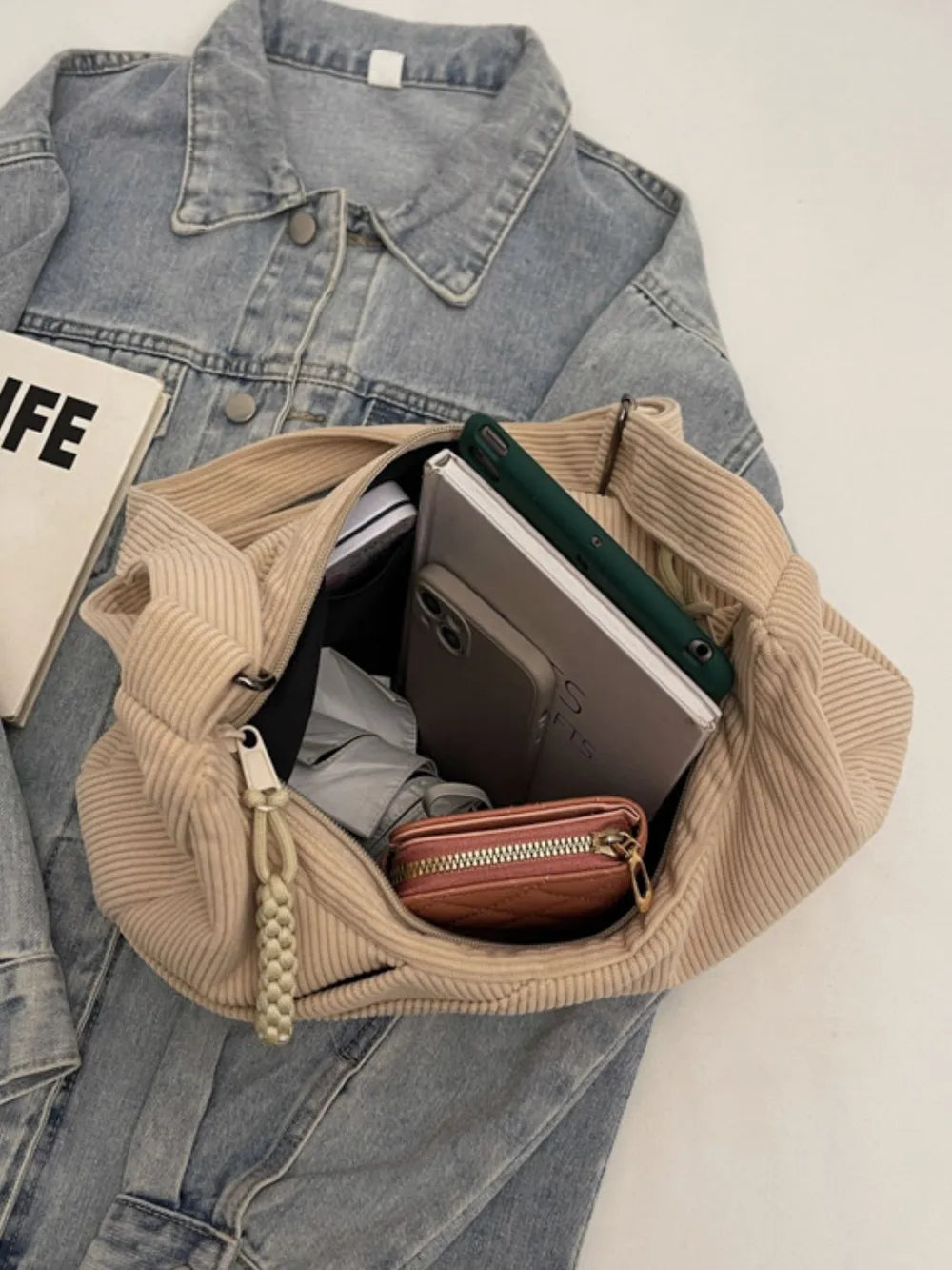 Beige corduroy crescent shoulder bag with braided tassel detail, shown with contents.