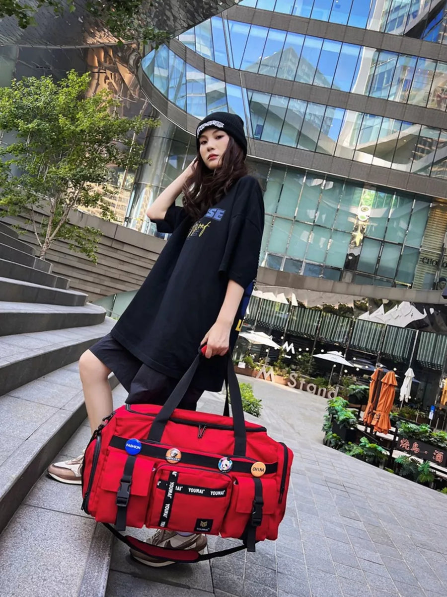 Red duffel bag with multiple pockets and decorative pins, held by a person wearing a black beanie and oversized t-shirt.