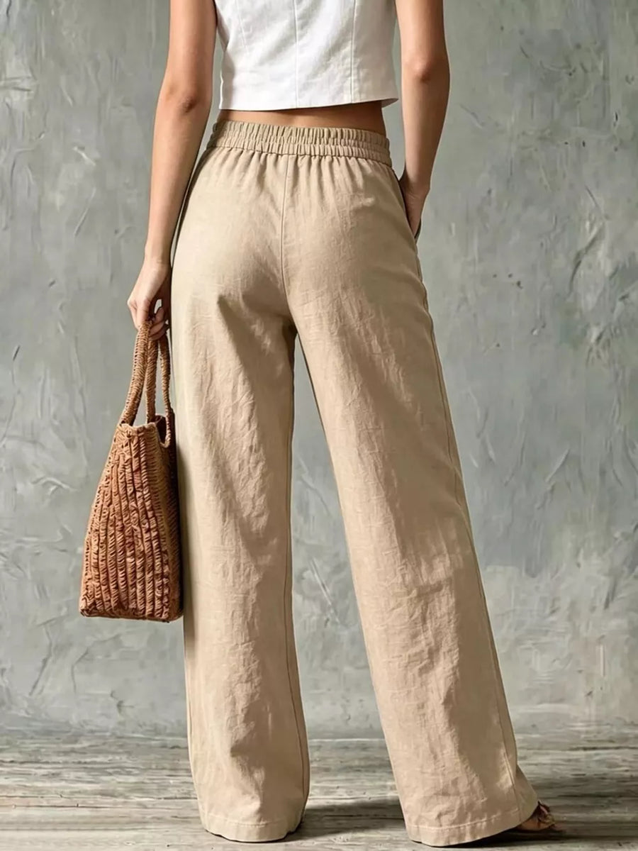 Beige linen wide-leg pants with elastic waistband and side pockets, styled with a straw bag.