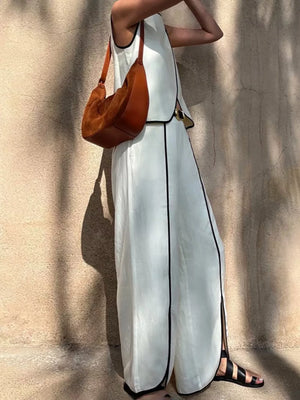 White sleeveless maxi dress with black trim and side slits, styled with a brown suede shoulder bag.