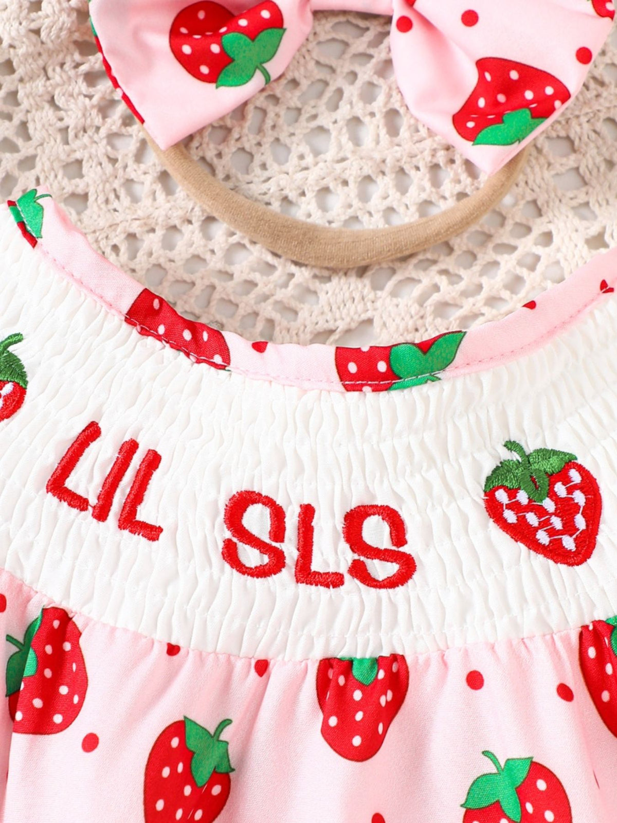 Baby one-piece in blush pink with strawberry print, smocked bodice, and embroidered text.