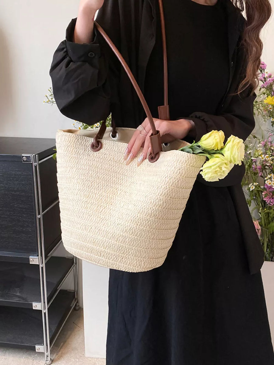 Natural straw woven large shoulder bag with brown leather straps, holding yellow flowers.