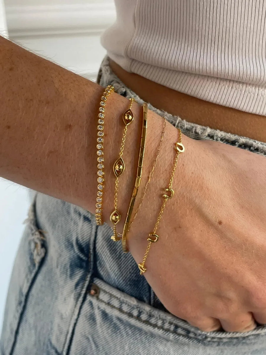 Gold charm chain bracelets with evil eye, bar, and letter charms.
