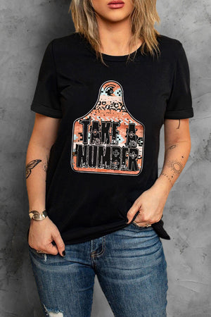 Black graphic t-shirt with "Take A Number" text on a cow tag shaped graphic with leopard print and pixelated pattern.