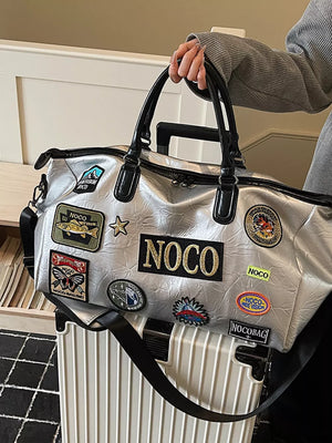 Silver metallic travel bag with textured finish and embroidered patches, featuring black handles and strap.