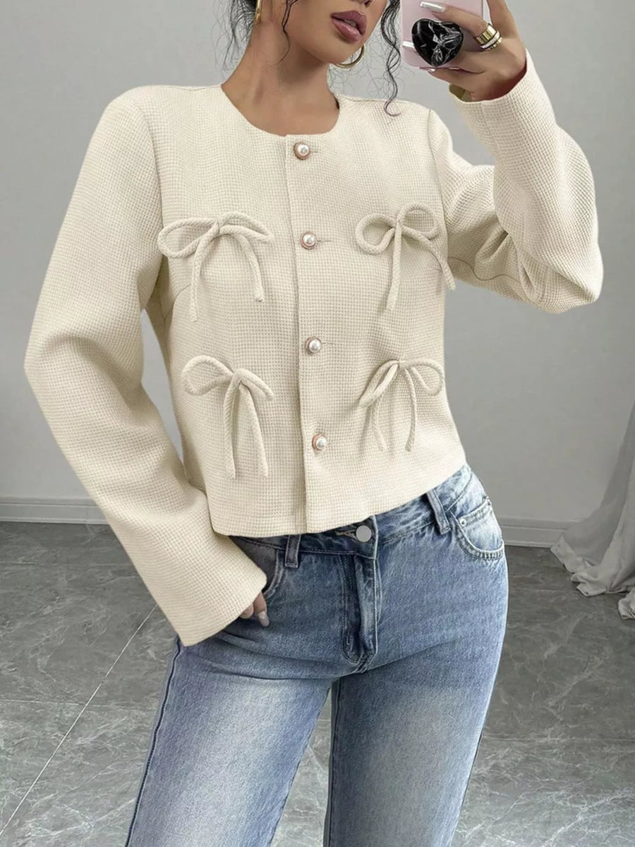 Cream textured jacket with round neck, pearl buttons, and bow details.