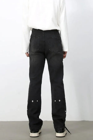 Dark wash denim trousers with asymmetric seams and decorative grommets on the lower legs.