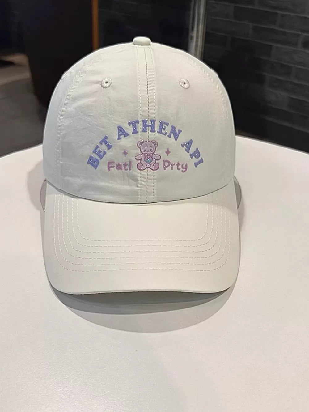 White baseball cap with purple embroidered text and a teddy bear graphic.
