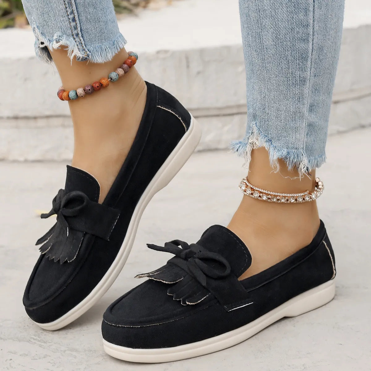 Black suede loafer flats with fringe and bow detail, styled with distressed jeans.