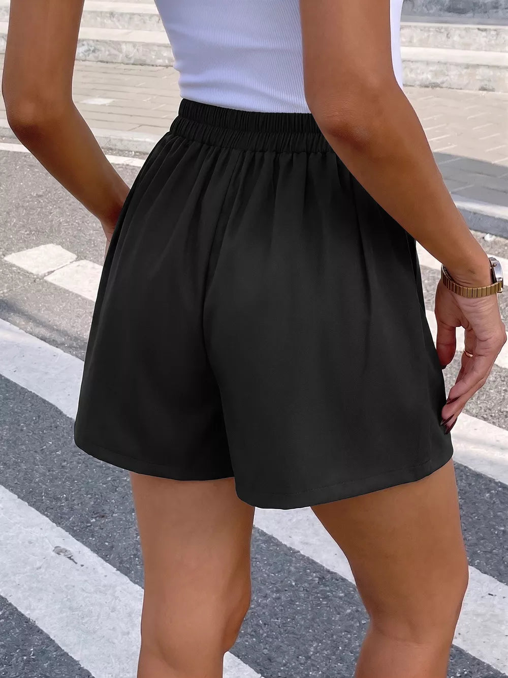 Black high-waisted pleated chino shorts with elastic waistband and decorative button detail.