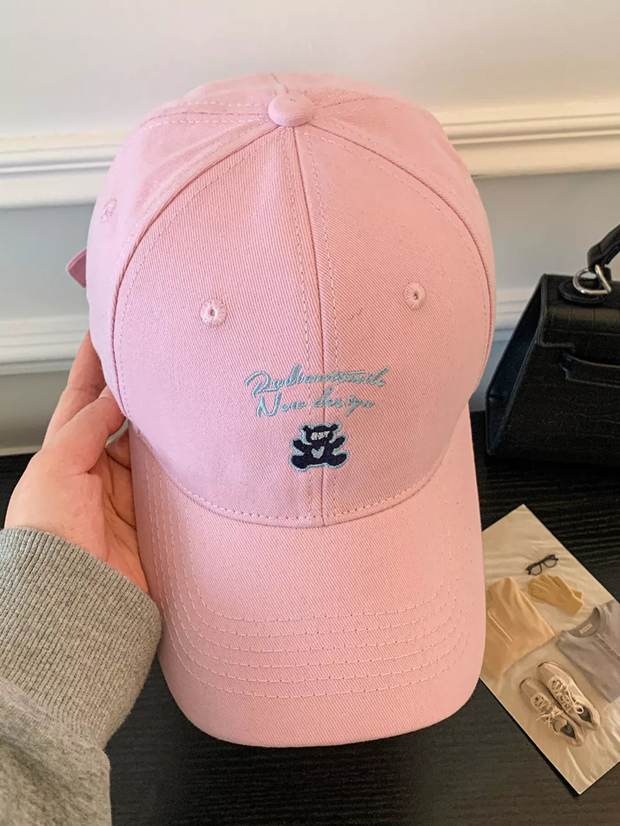 Pink baseball cap with embroidered teddy bear and script text detail.