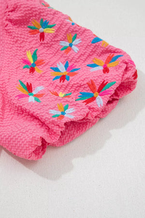 Women's pink textured blouse with colorful embroidered floral details on the sleeve.
