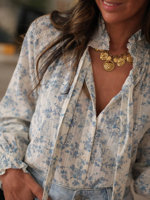 White long-sleeve blouse with blue floral print, tie neck, and ruffled cuffs.