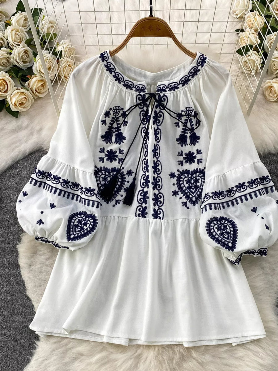 White tie-neck blouse with intricate blue embroidery and balloon sleeves.
