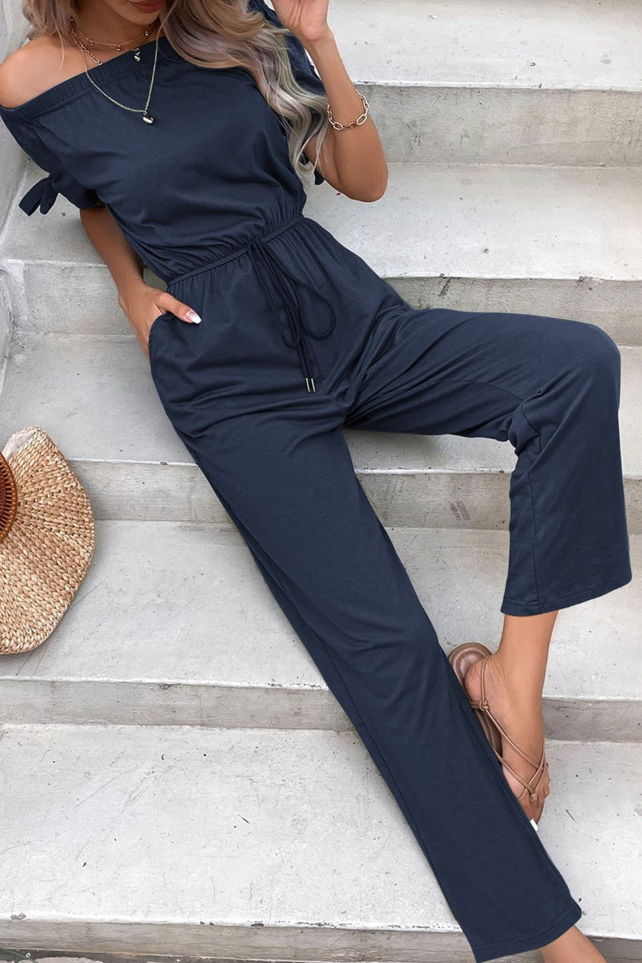 Off-the-shoulder navy jumpsuit with tie sleeves, drawstring waist, and pockets.