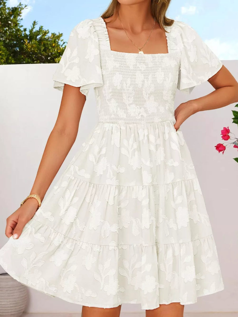 White smocked mini dress with tiered skirt, flutter sleeves, and floral embroidery.