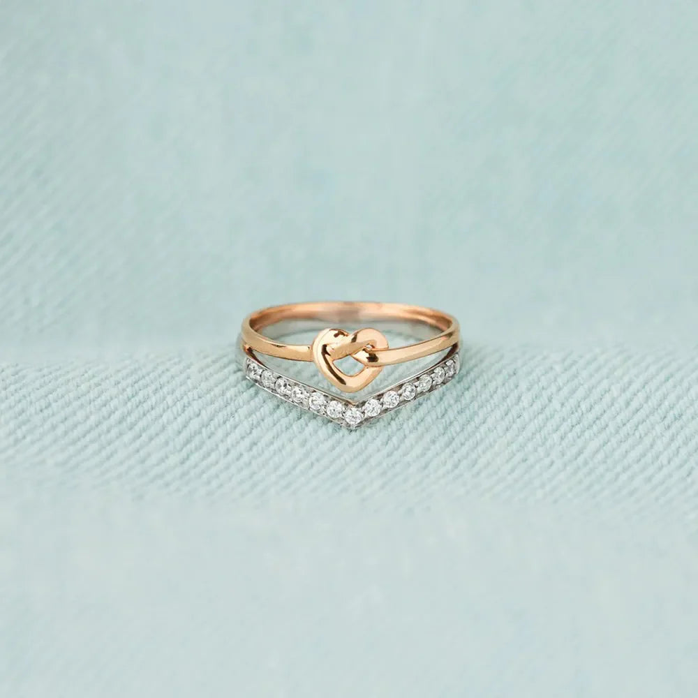 Stacking rings with a gold heart knot and a chevron band of cubic zirconia on a light blue textured background.