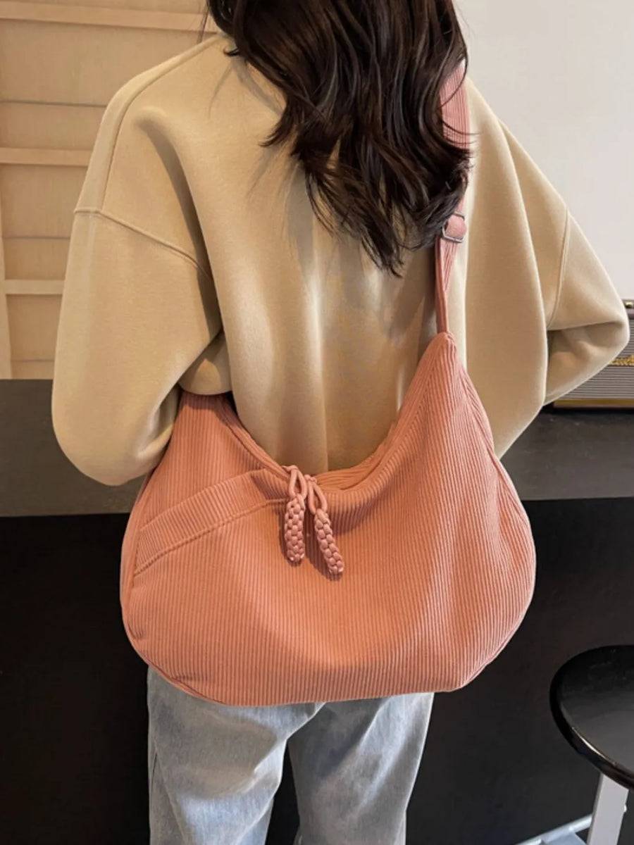 Pink corduroy crescent shoulder bag with braided tassels and front pocket.