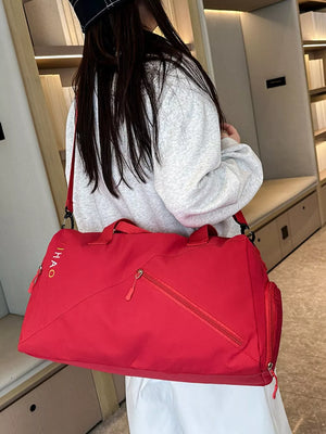 Red duffel bag with diagonal zip pocket and side pouch, embroidered logo detail.