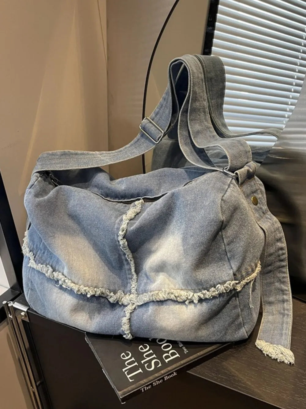 Light wash denim slouch crossbody bag with frayed edges and adjustable strap.