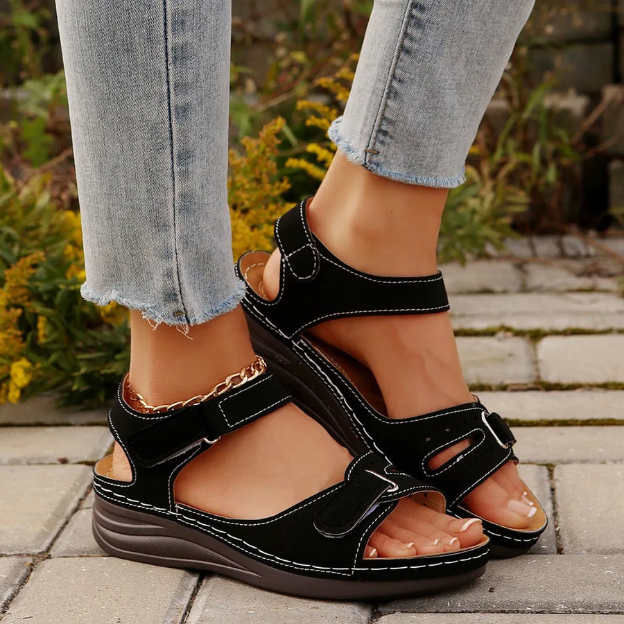 Black perforated velcro wedge sandals with white stitching and a gold chain ankle strap.