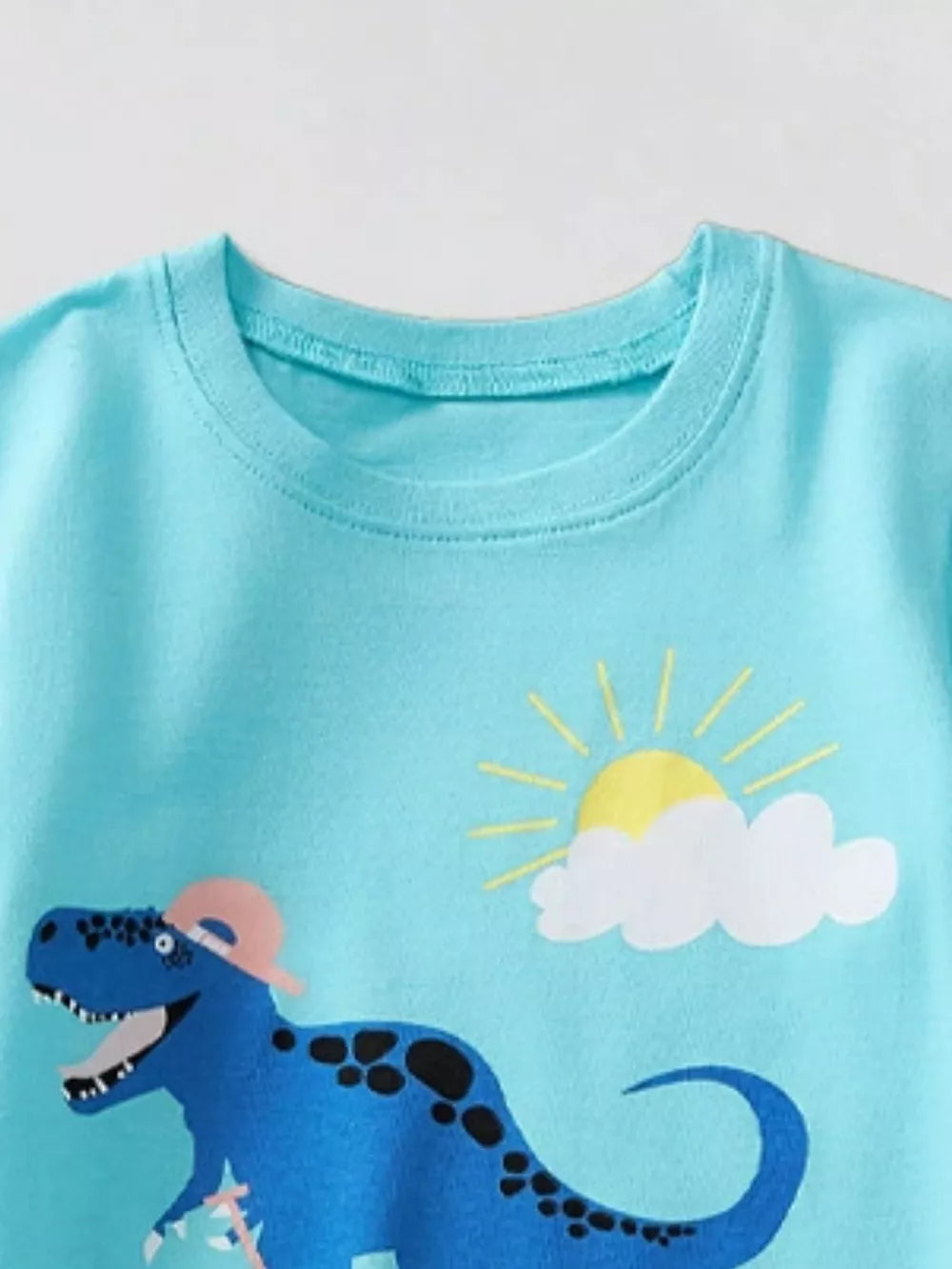 Kids blue t-shirt with a graphic of a blue dinosaur wearing a pink hat and a sun with clouds.