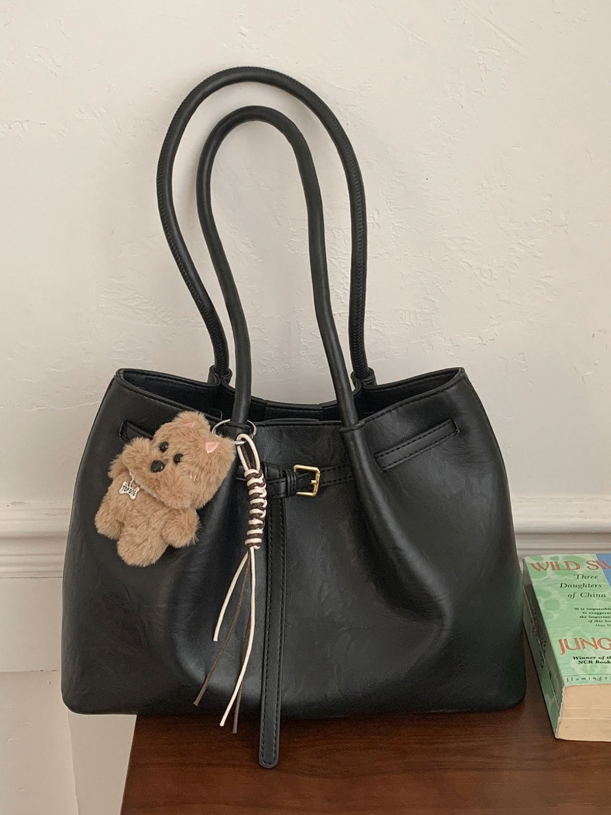Black leather shoulder bag with buckle detail, braided tassel, and teddy bear charm