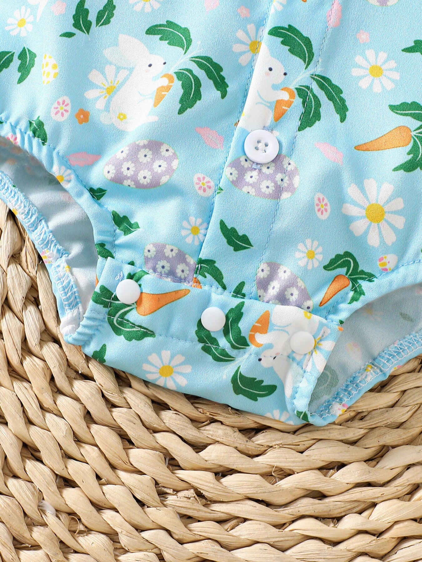 Baby bodysuit with bunny, carrot, and flower print on blue fabric, featuring white buttons.